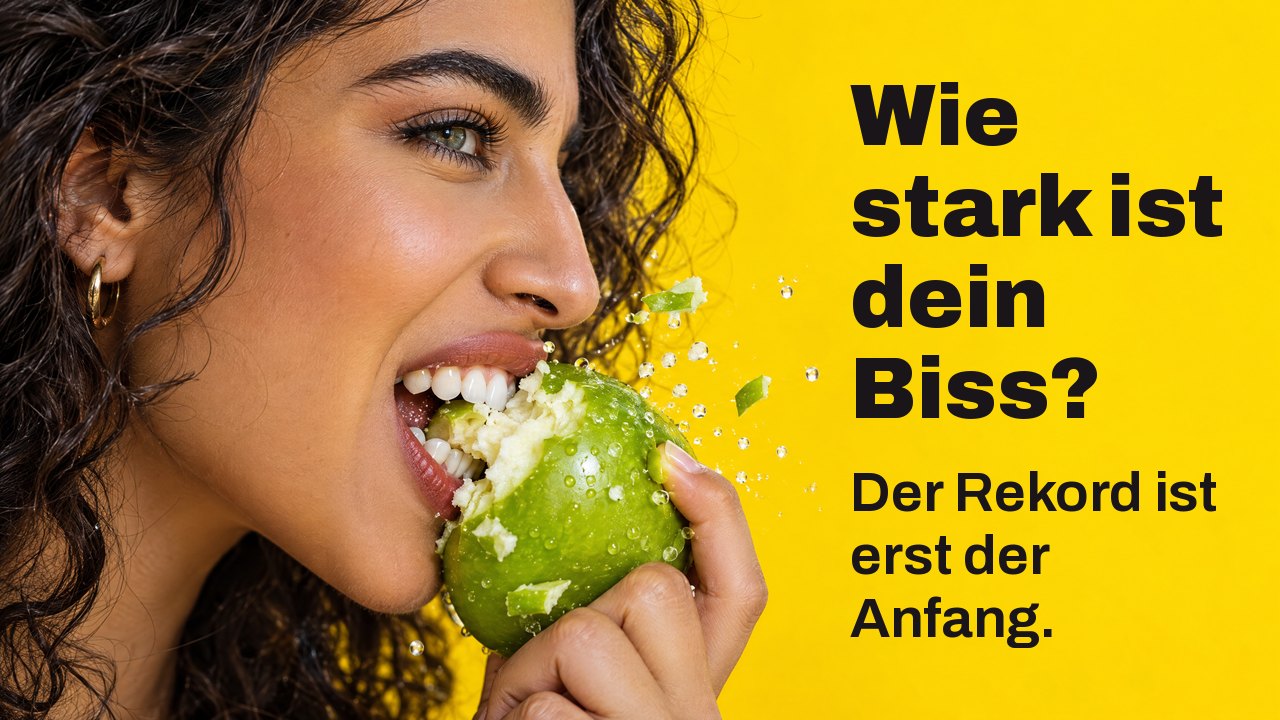 Wie stark ist dein Biss? Der Rekord ist erst der Anfang. Nahaufnahme im Profil: Eine Frau beißt kräftig in einen knackigen grünen Apfel, Stücke fliegen, vor leuchtendem Gelb.