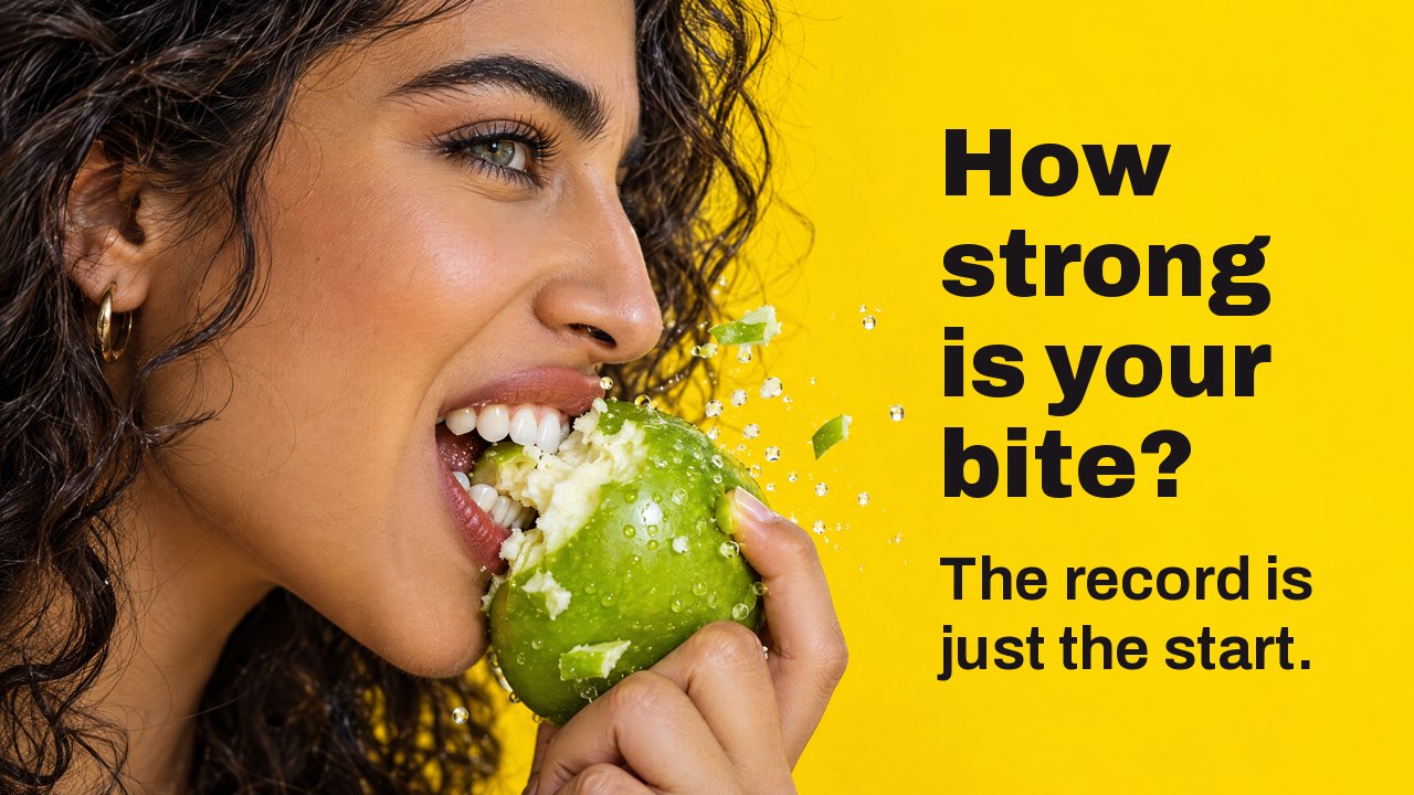 How strong is your bite? The record is just the start. Side profile close up of a woman biting hard into a crisp green apple, shards flying, against bright yellow.