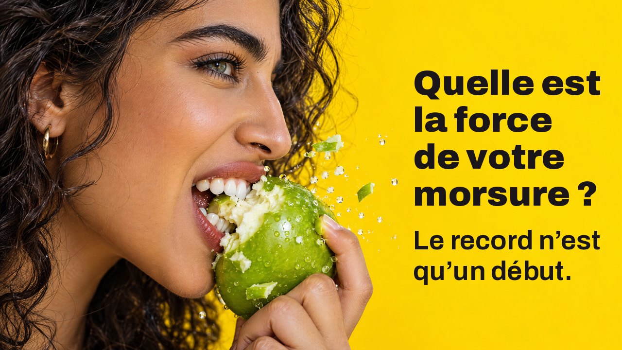 Quelle est la force de votre morsure ? Le record n’est qu’un début. Gros plan de profil d’une femme qui croque à pleines dents une pomme verte, des éclats volent, sur fond jaune vif.