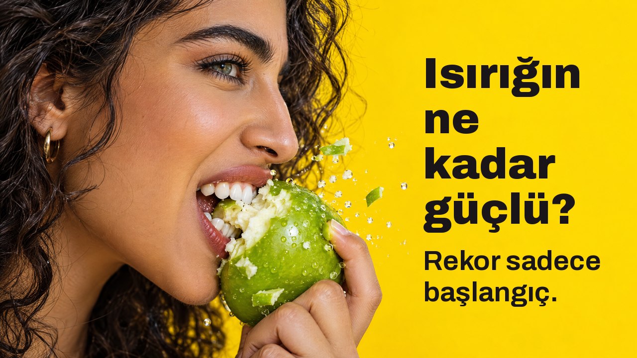 Isırığın ne kadar güçlü? Rekor sadece başlangıç. Yandan yakın çekim: Bir kadın gevrek yeşil elmayı sertçe ısırıyor, parçalar uçuşuyor, parlak sarı fon.