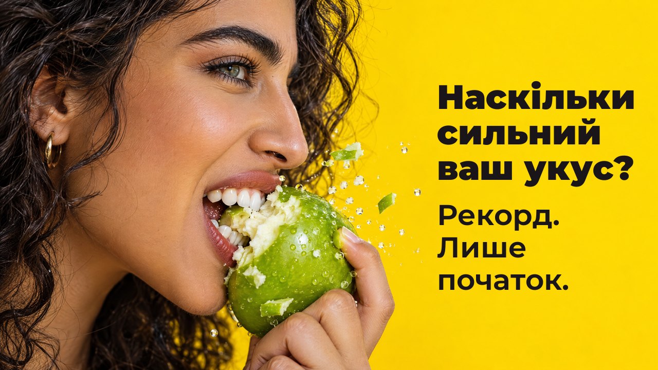 Наскільки сильний ваш укус? Рекорд: лише початок. Жінка в профіль великим планом з силою відкушує хрустке зелене яблуко, летять шматочки, на яскраво жовтому тлі.