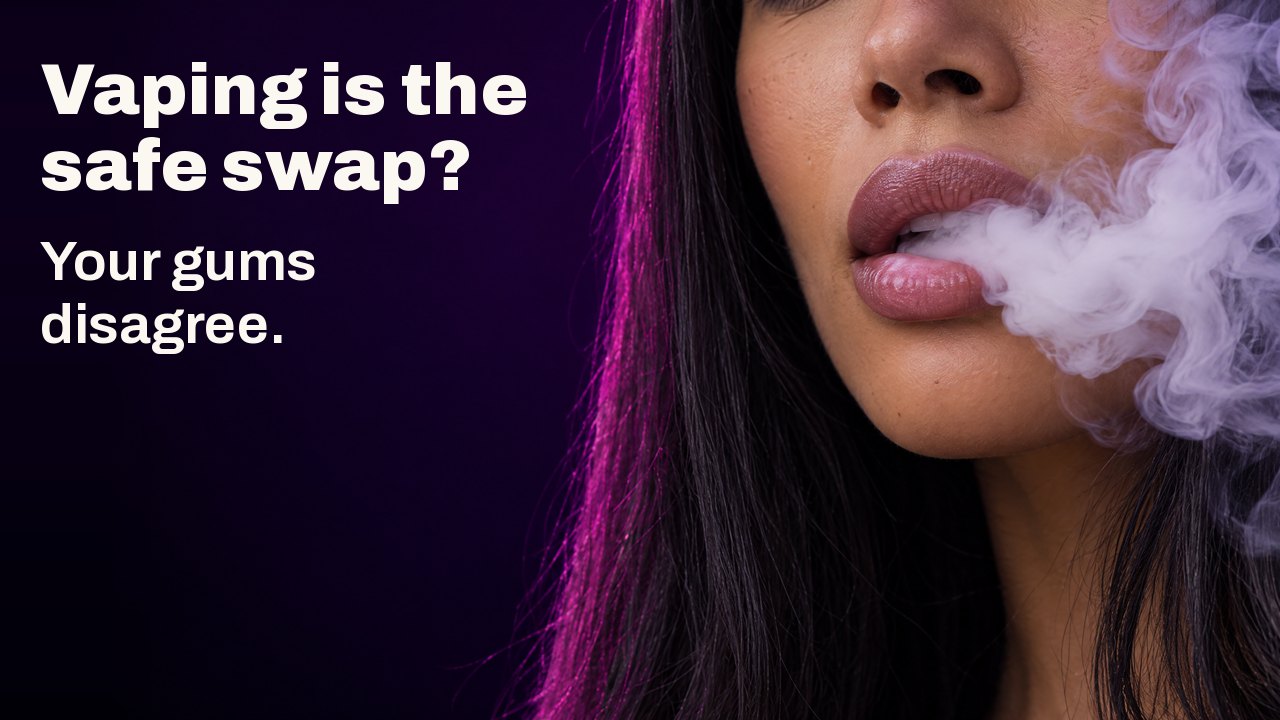 Vaping is the safe swap? Your gums disagree. Close up of a woman’s lips exhaling a thick swirling cloud of white vapour against a dark violet background.